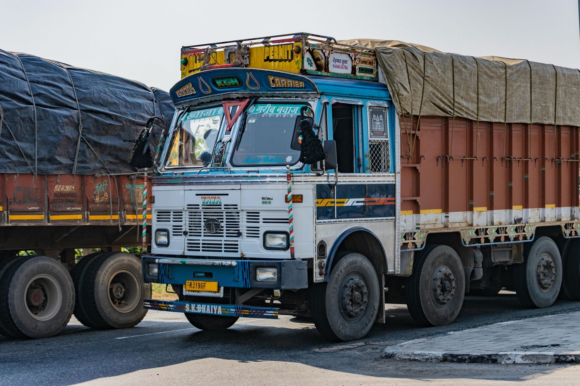 A white and blue Tata truck loaded with cargo travels on a road alongside another truck under a clear, bright sky.