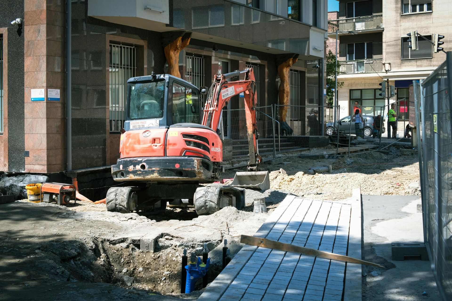 An orange excavator operates on a construction site outside a modern office building, with a pit in the foreground.