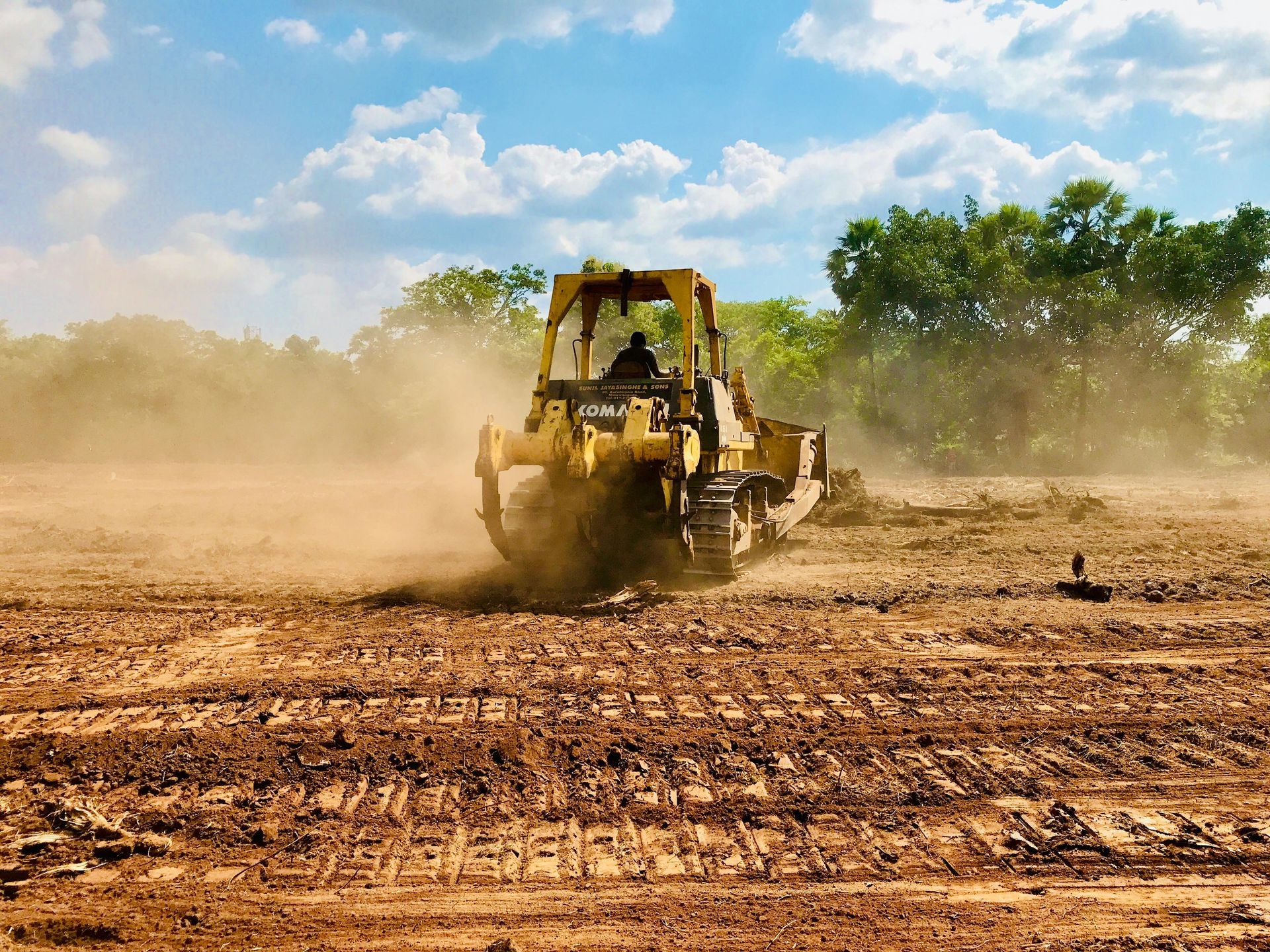 A yellow bulldozer pushes dirt across a sunny, dusty construction site with trees in the background.