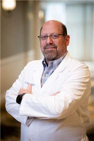 A person wearing a white lab coat and glasses standing with arms crossed in an office setting.