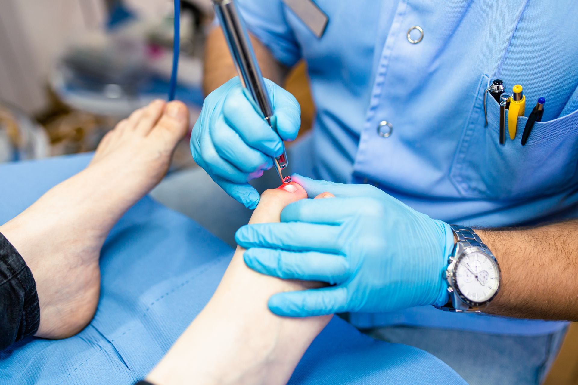 A podiatrist in blue gloves examines a patient's foot with surgical tools; setting appears to be a doctor's office.
