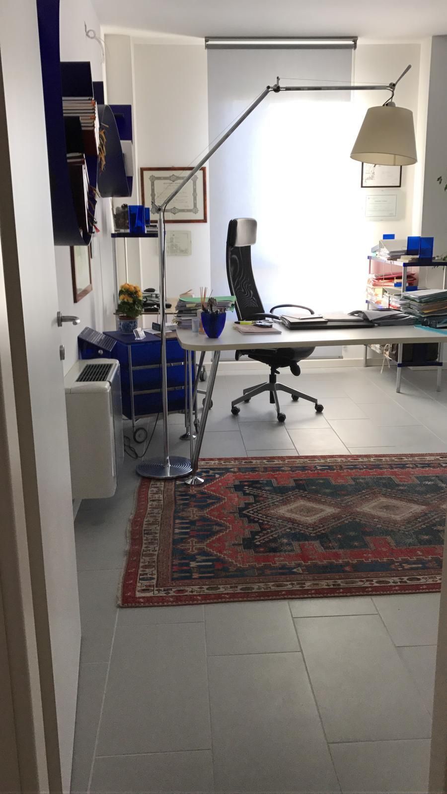 Ufficio con scrivania, sedia, lampada e tappeto. Sulla sinistra, librerie e un termosifone. La luce filtra da una finestra.