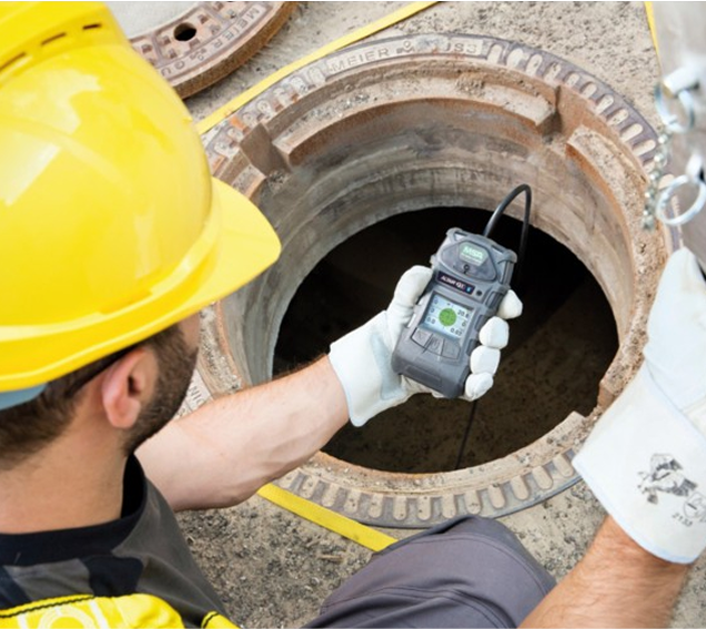 A man wearing a hard hat and gloves is holding a device in front of a manhole cover