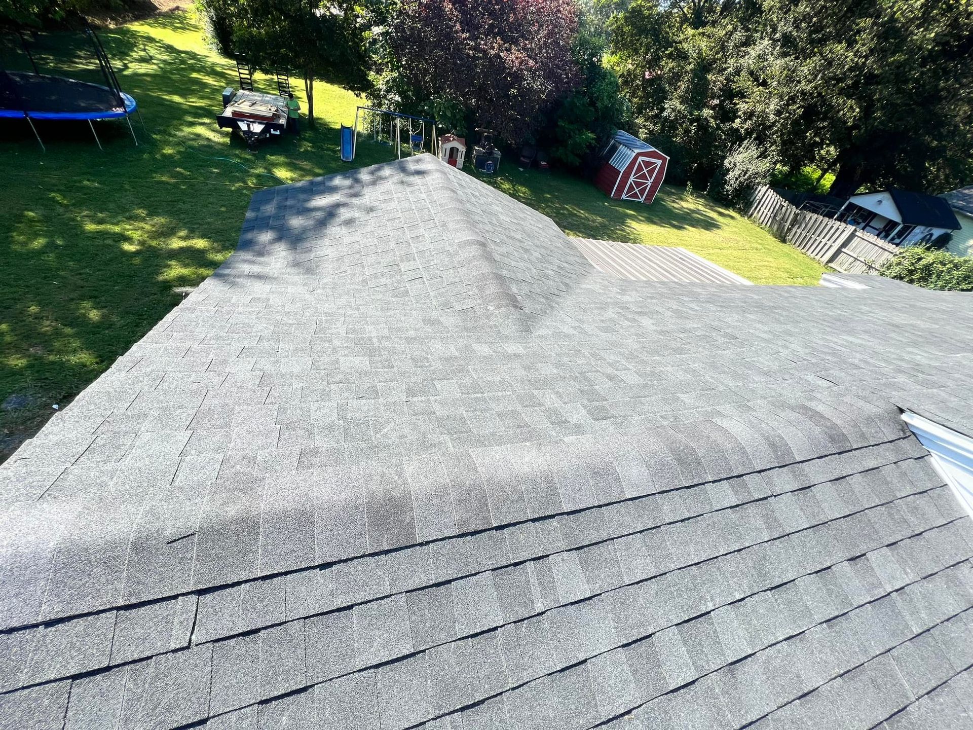 Gray asphalt shingle roof on a house, angled view. Green lawn, trees, and small buildings in background.