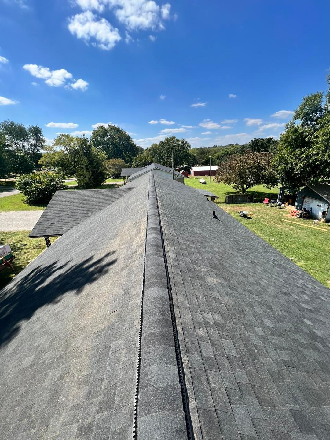 A dark-gray shingled roof under a blue sky, viewed from a high angle. Green trees and grass are in the background.