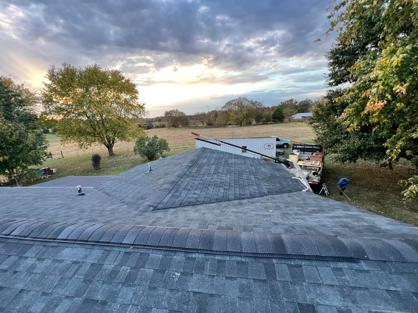 Overhead view of a house roof, cloudy sky. Workers are visible on the ground near a truck in a field.