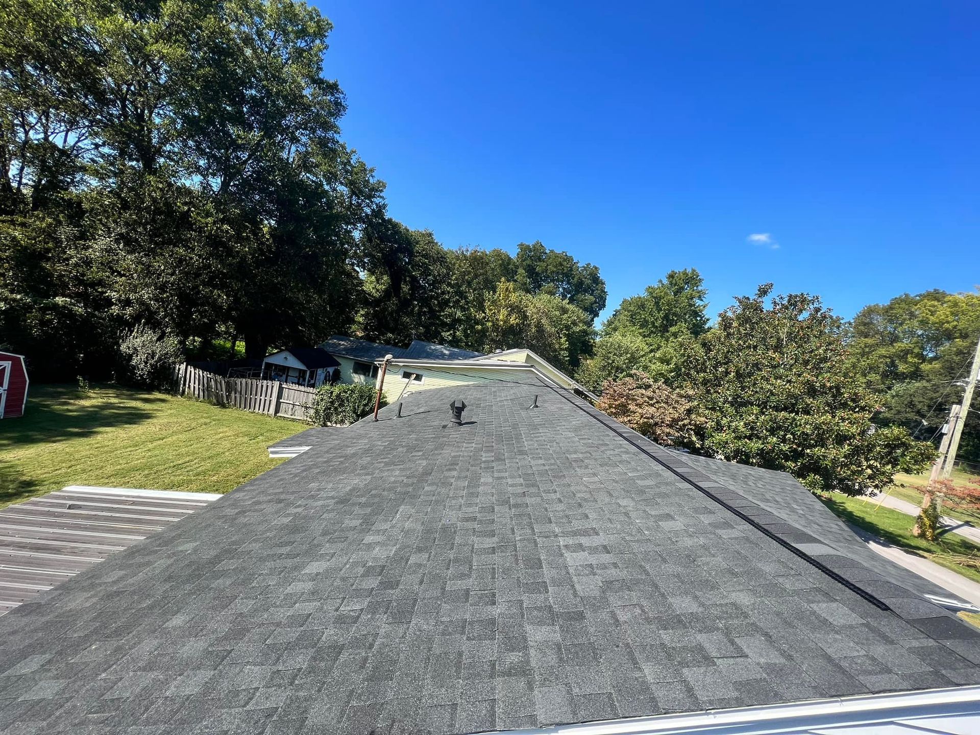 A dark gray shingled roof under a clear blue sky, with trees in the background.