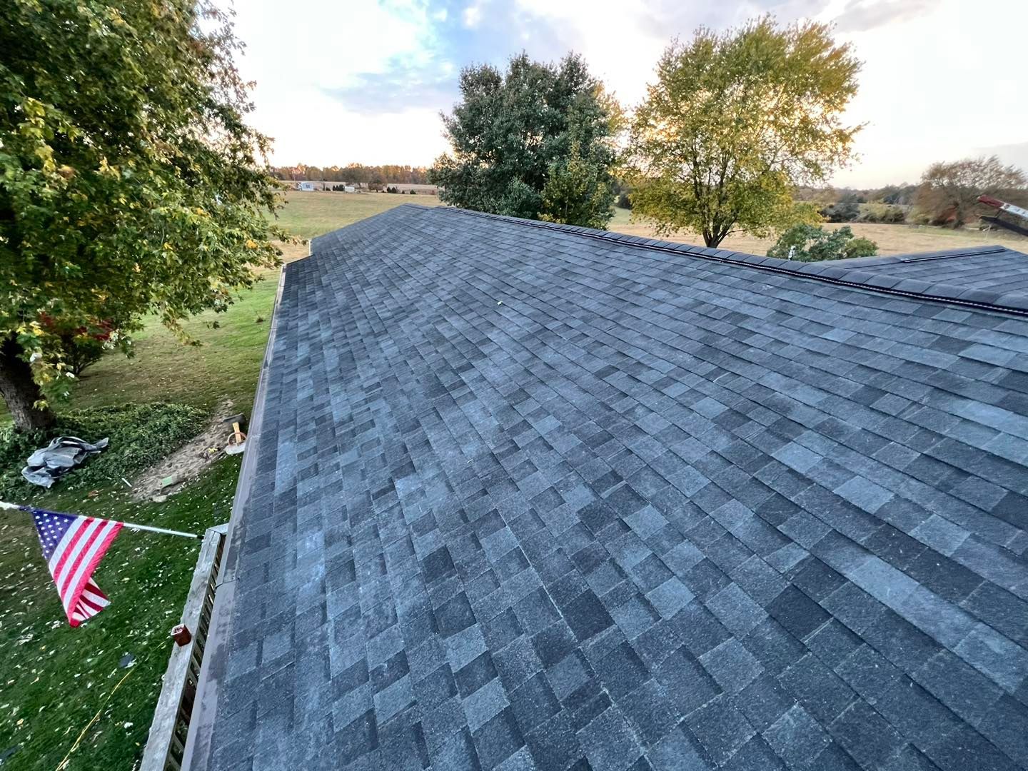 Dark gray asphalt shingle roof on a house, American flag waving in the breeze, trees and sky in the background.