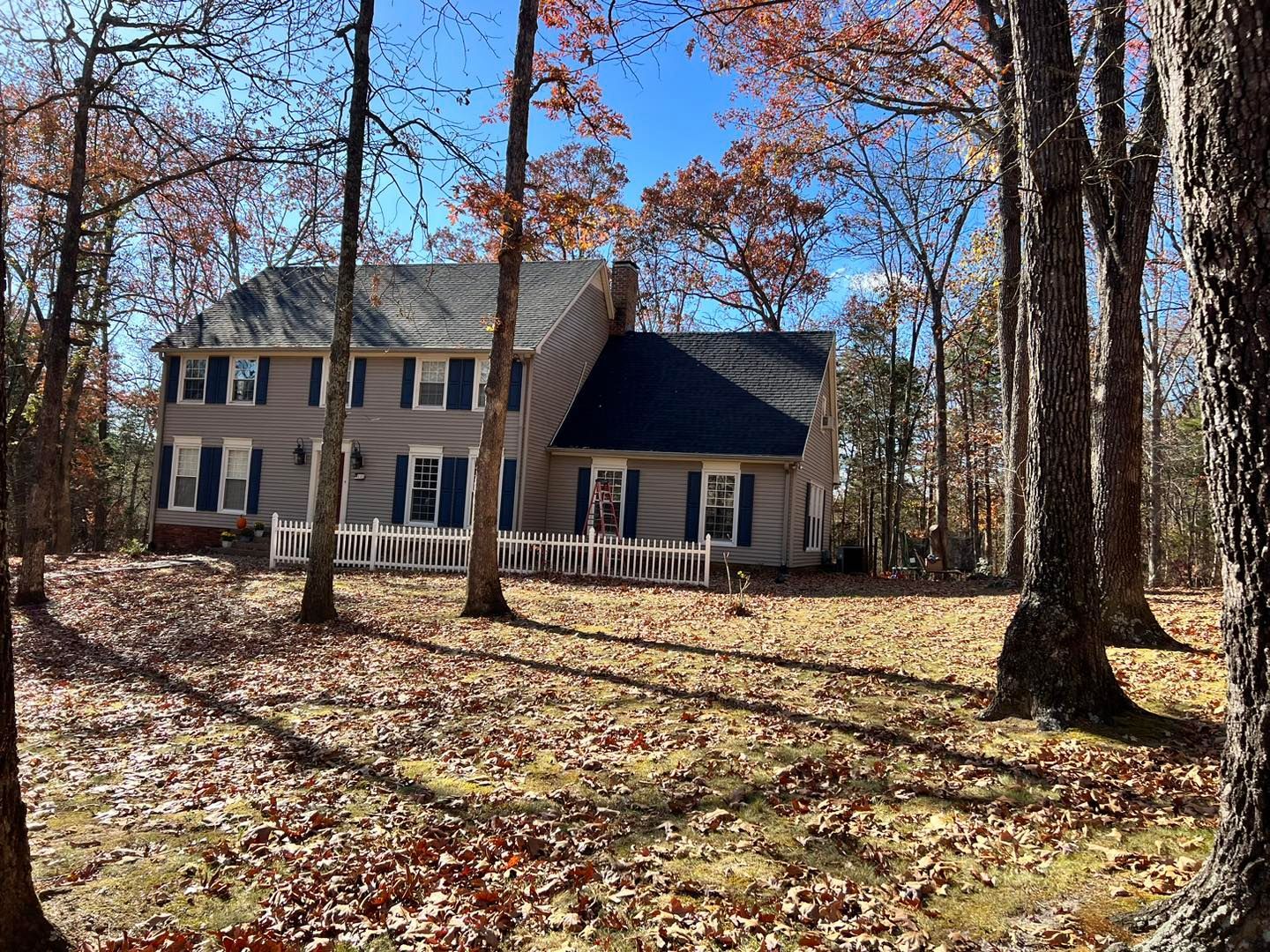 Two-story house with blue shutters, a white picket fence, and autumn trees on a sunny day.