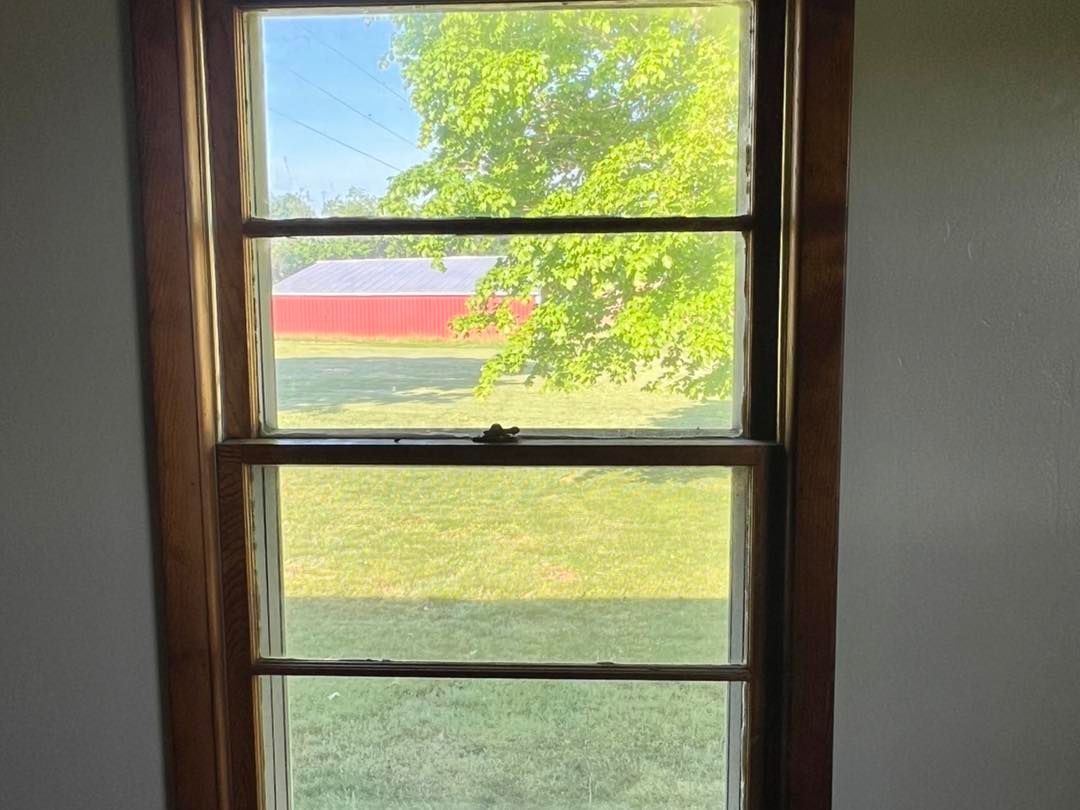 Wooden-framed window looking out onto a green yard with a red barn and tree in the distance.