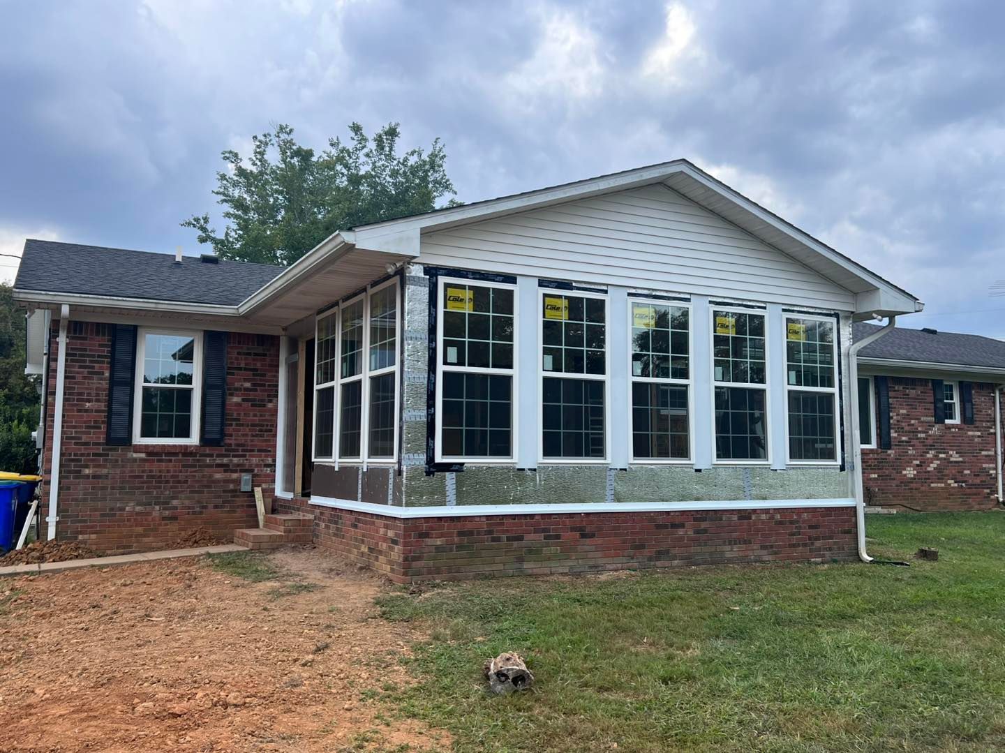 Brick house with sunroom addition, new windows, and unfinished siding. Overcast sky.