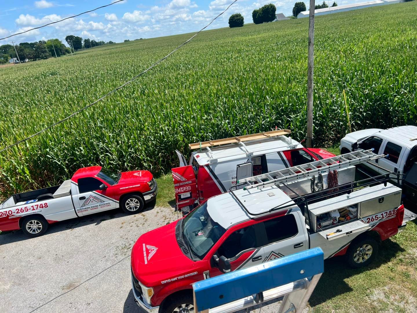 Red and white work trucks parked near a green cornfield on a sunny day.