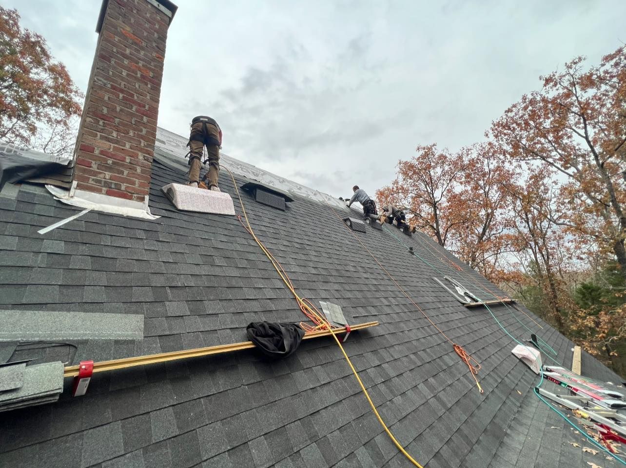 Roofers working on a dark gray shingled roof, next to a brick chimney and trees.
