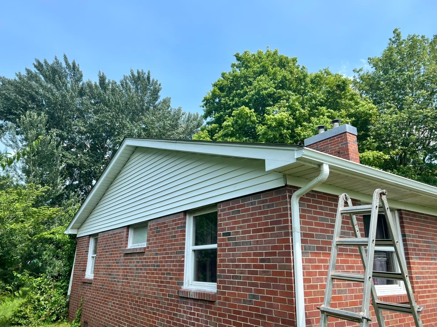 Brick house with white siding and gutters, surrounded by trees, blue sky. A ladder is propped against the roof.