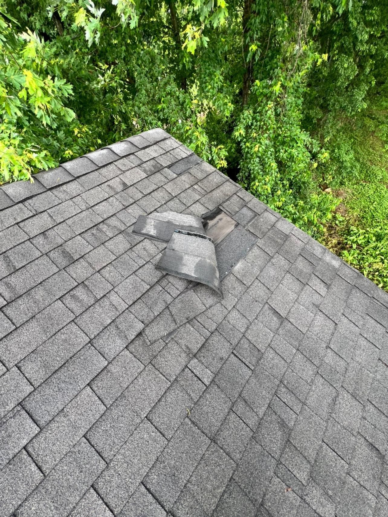 Damaged asphalt shingle roof, top view with missing and broken shingles; green trees in background.