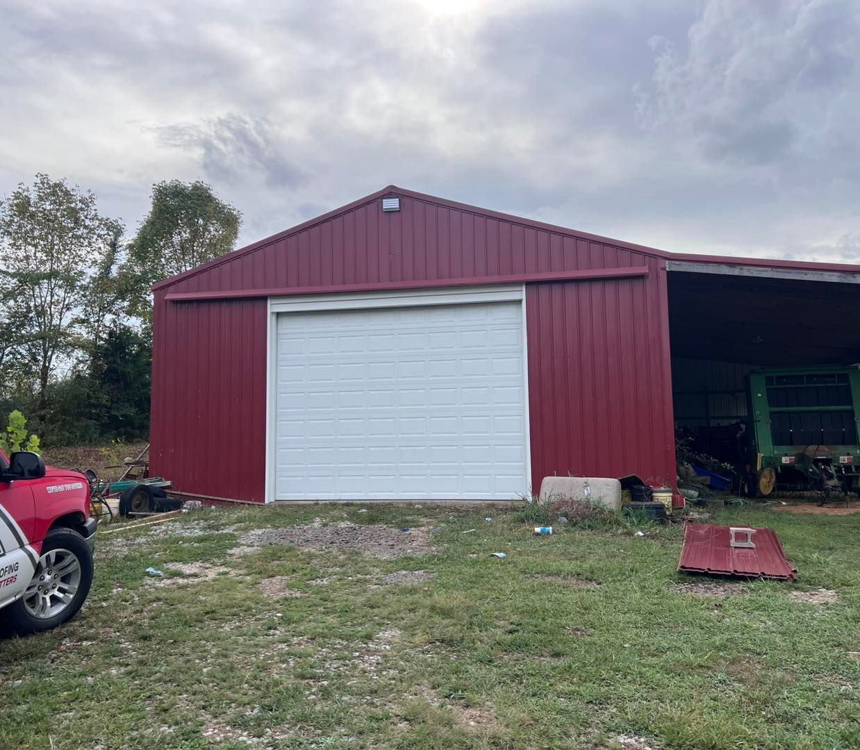 Red metal barn with white garage door and open side, set in a grassy yard under a cloudy sky.