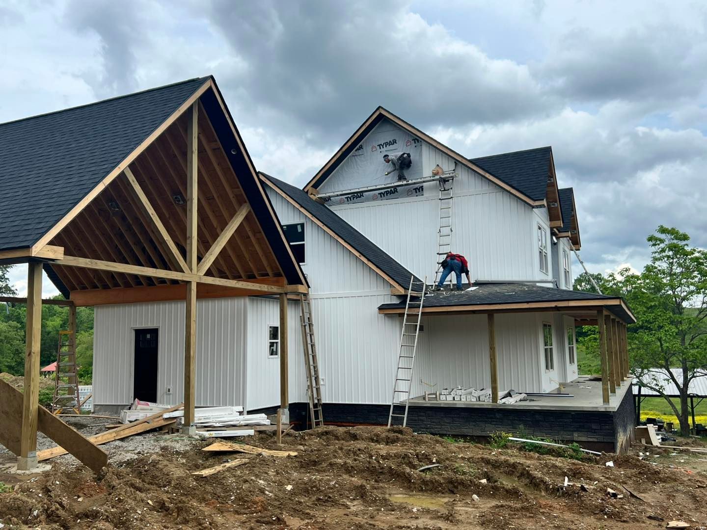 House under construction, white siding, black roof, wood framing, worker on roof, cloudy sky.