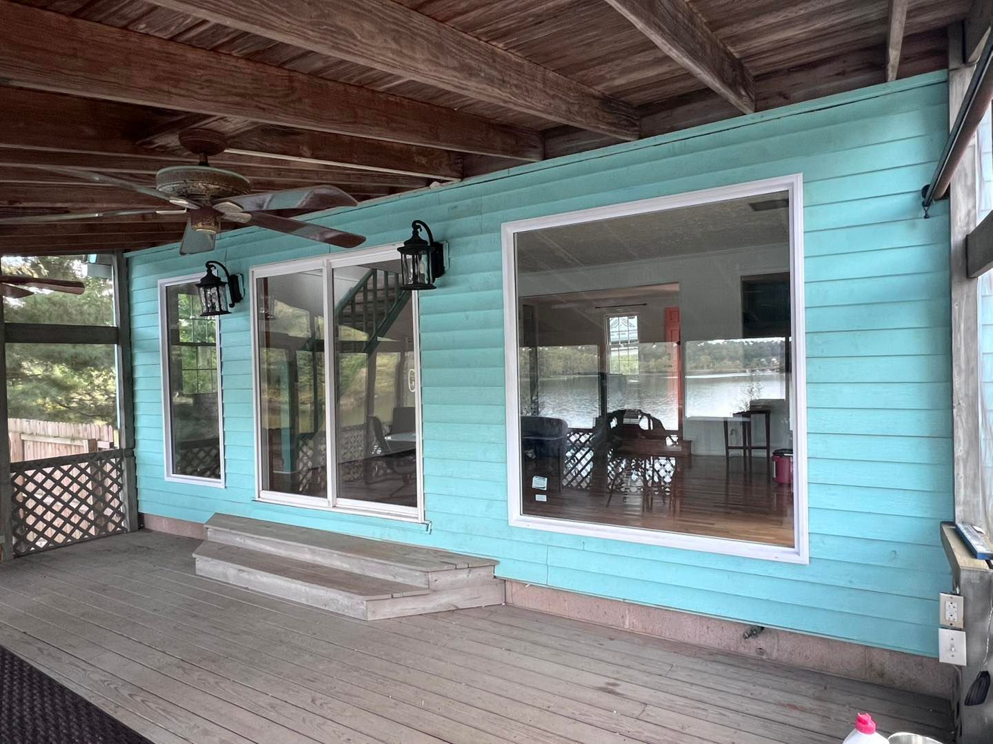 Covered porch with teal siding and large windows, reflecting interior. Wooden deck, stairs, and ceiling.