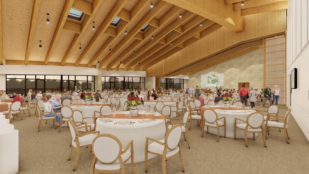 Large event space, round tables with white linens, guests seated, wood ceiling, large windows.