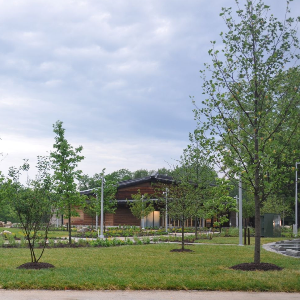 Curving paved path beside a grassy field and modern building at sunset, with trees and pink sky beyond.