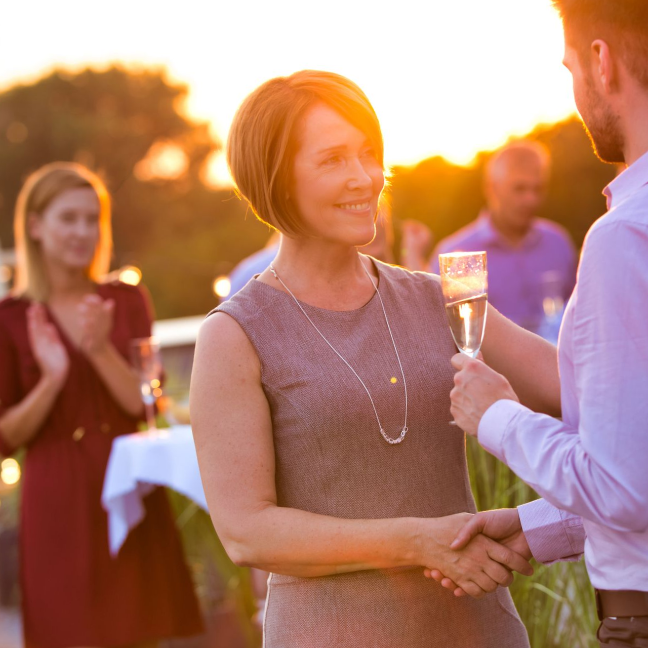Woman in gray dress shaking hands, holding champagne, at a party, golden hour lighting.