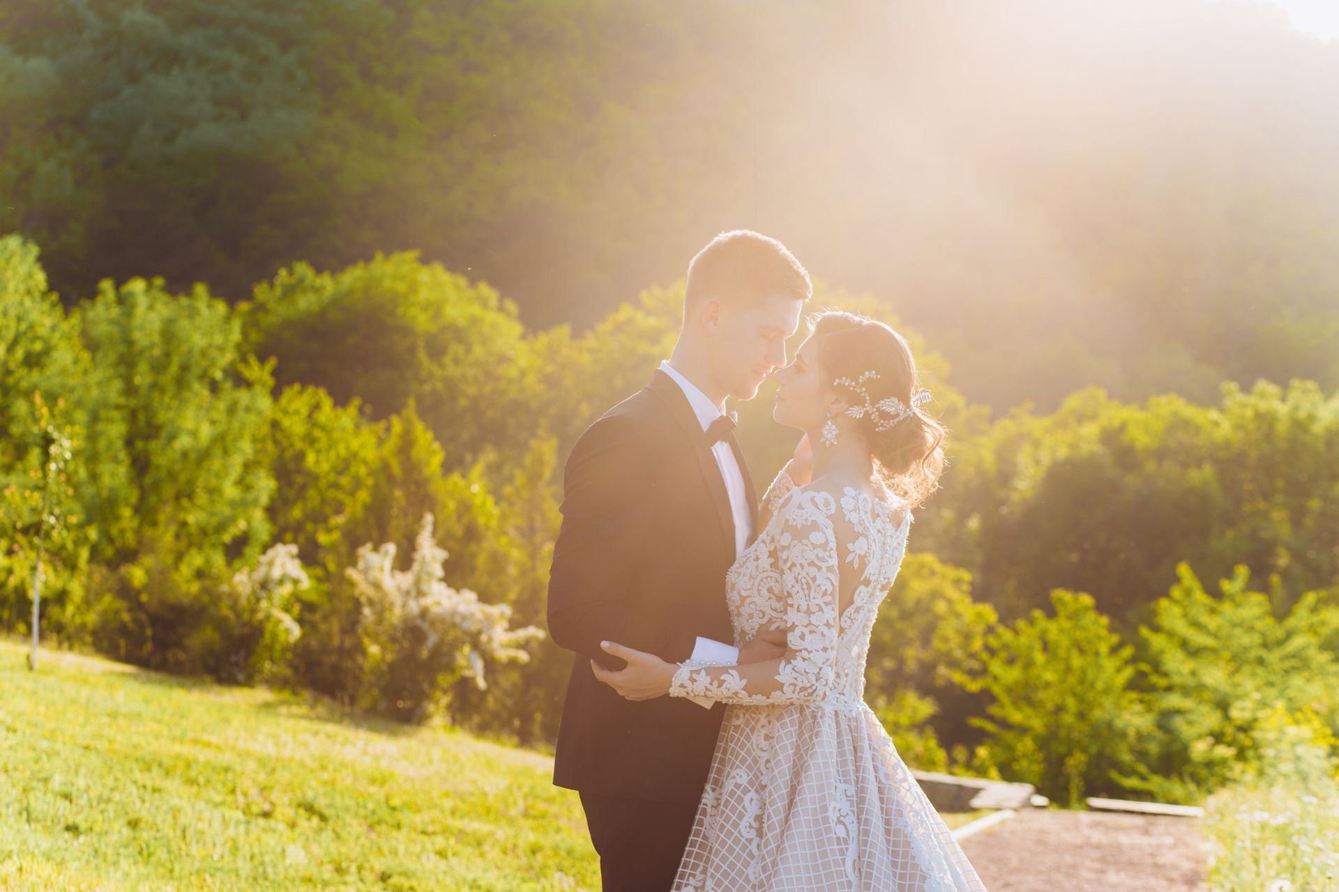 Couple kissing, bride in lace dress, groom in tuxedo,  park backdrop, bright sunlight.