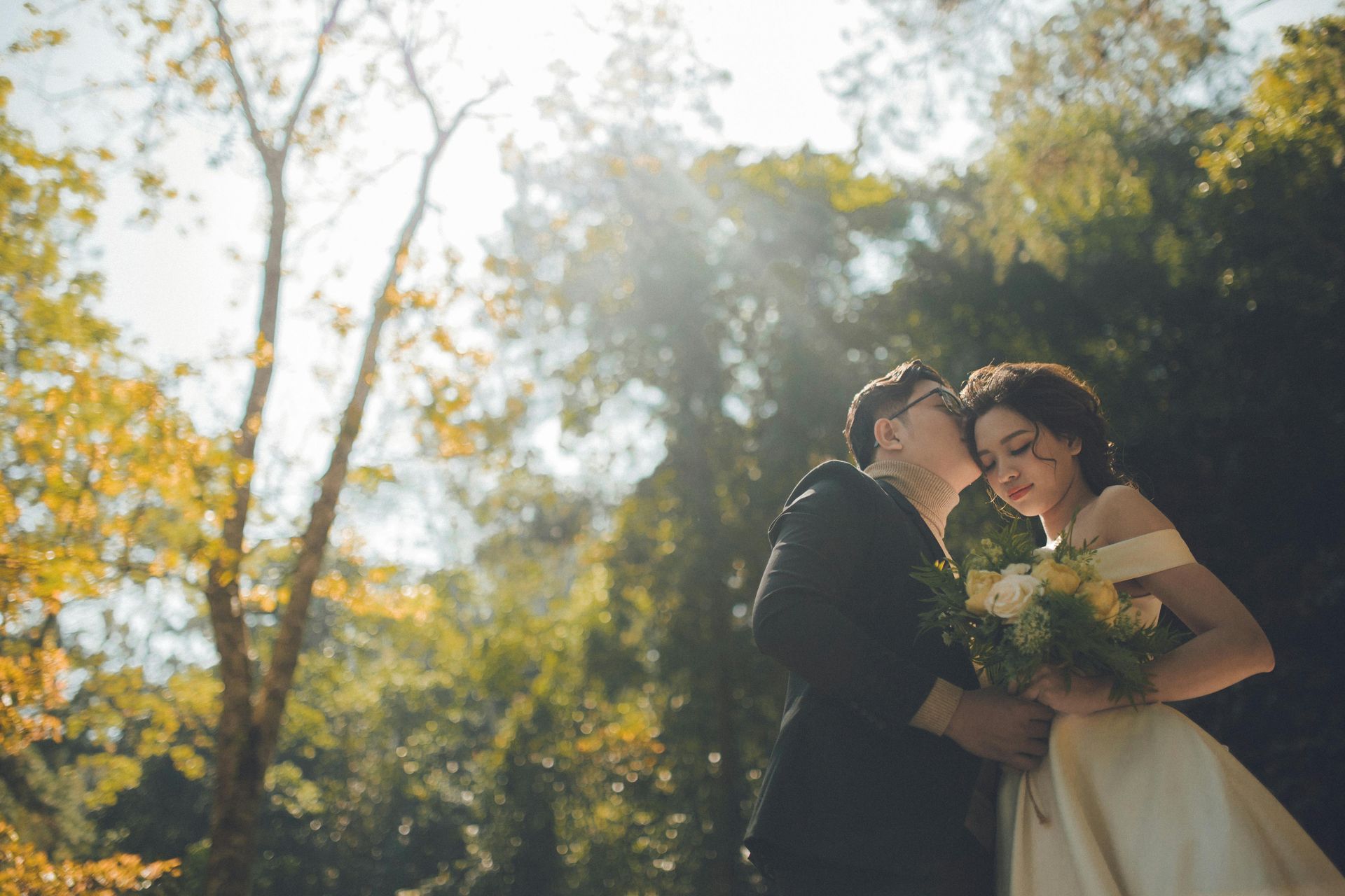 Couple embracing outdoors; the man kisses the woman's cheek, sunlight streams through trees in the park.