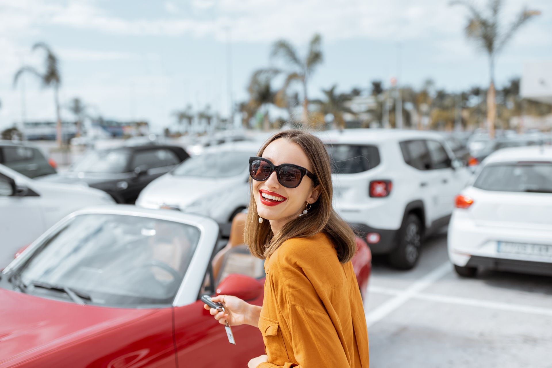 Portrait of a beautiful young woman standing with keys near the red cabriolet at the car parking outdoors.