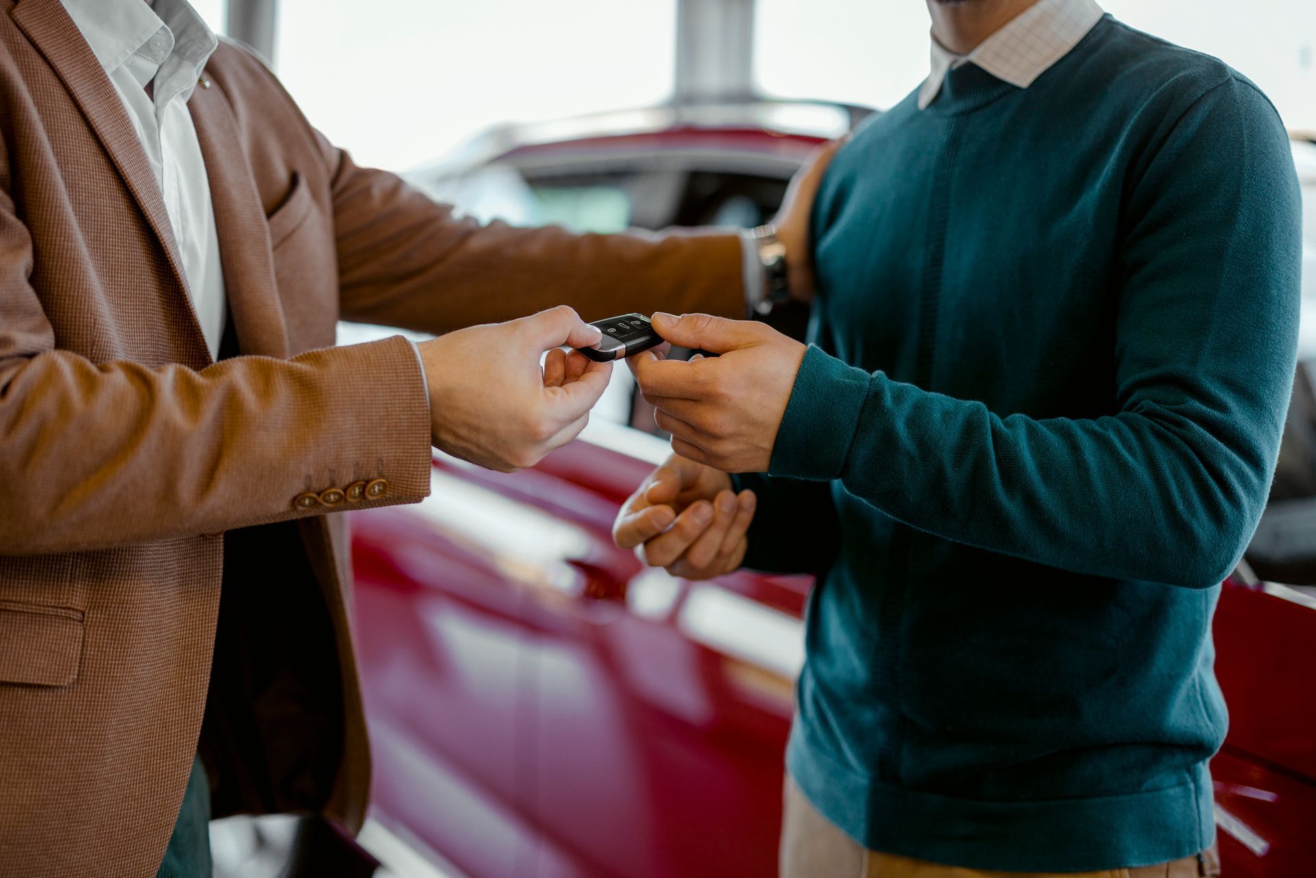 Two people exchange a car key in front of a red vehicle.