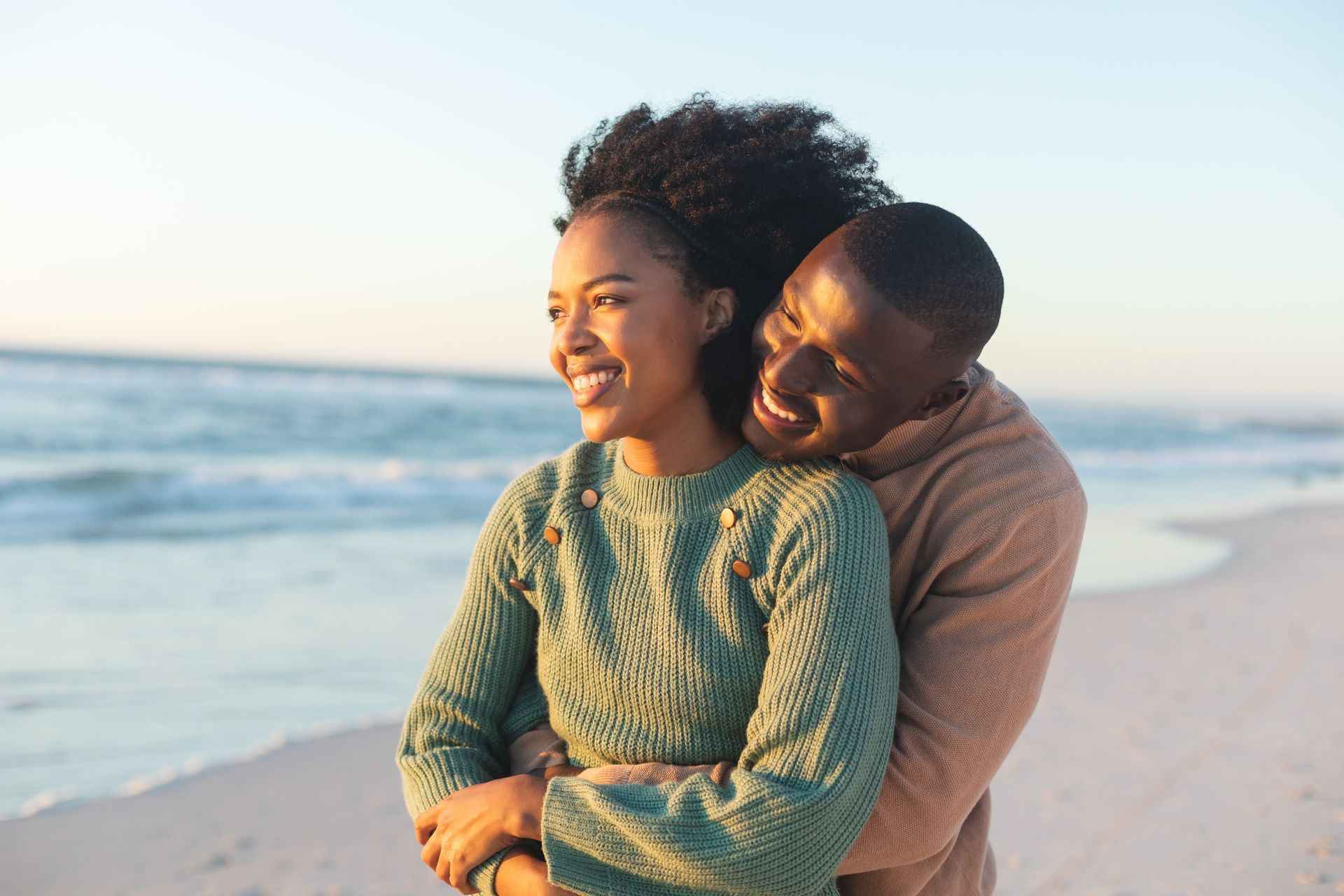 Smiling senior couple on a beach: Woman embraces man from behind, looking toward the sun.