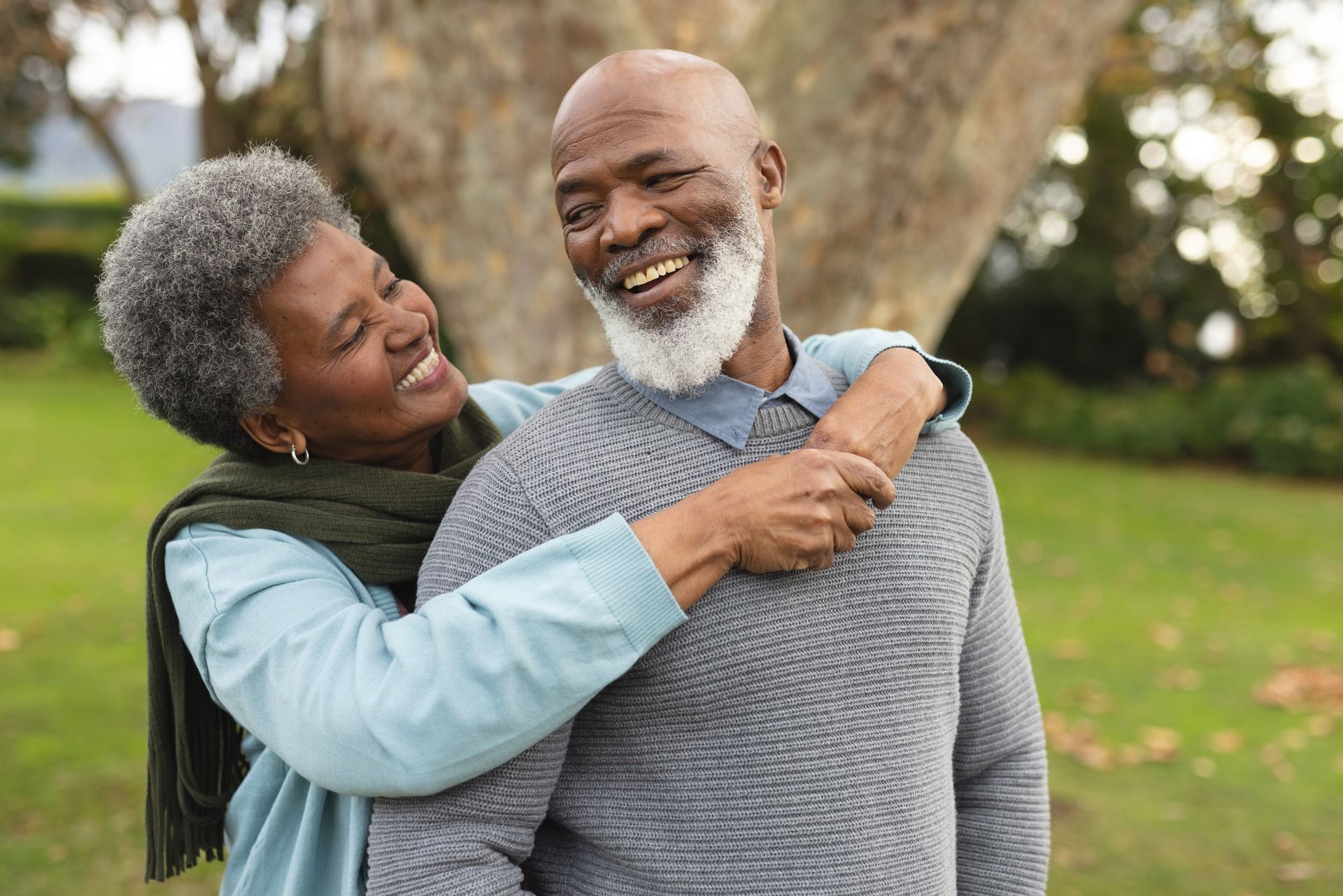 Smiling couple embracing outdoors. Woman with gray hair, arms around man’s shoulders.