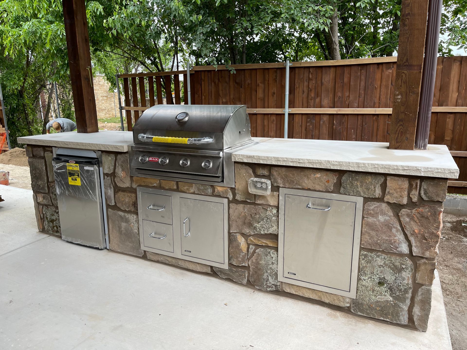 Outdoor patio with stone fireplace, grill, and wicker furniture under a sunny blue sky with pine trees in the background.