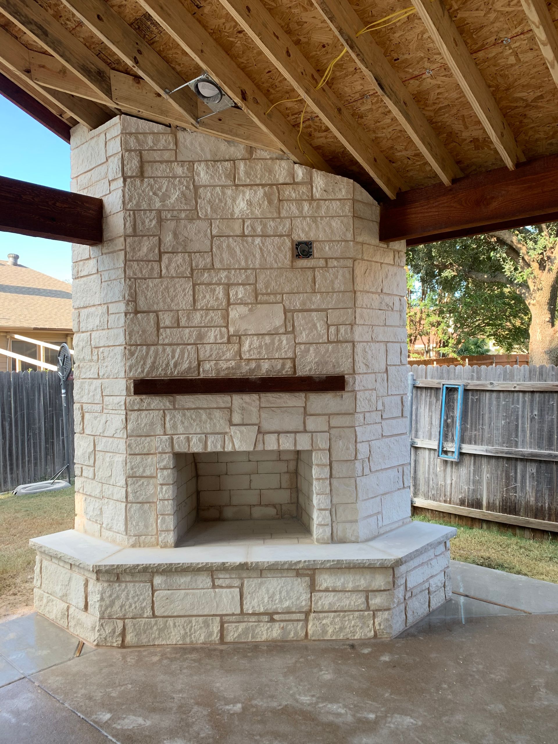 A stone outdoor fireplace with a burning fire, situated between stone benches on a paved patio.