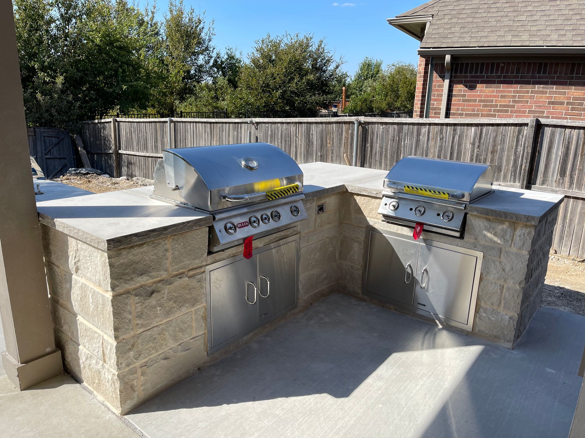 Modern outdoor kitchen with stone counters, wood cabinetry, and stainless steel appliances under a covered patio.