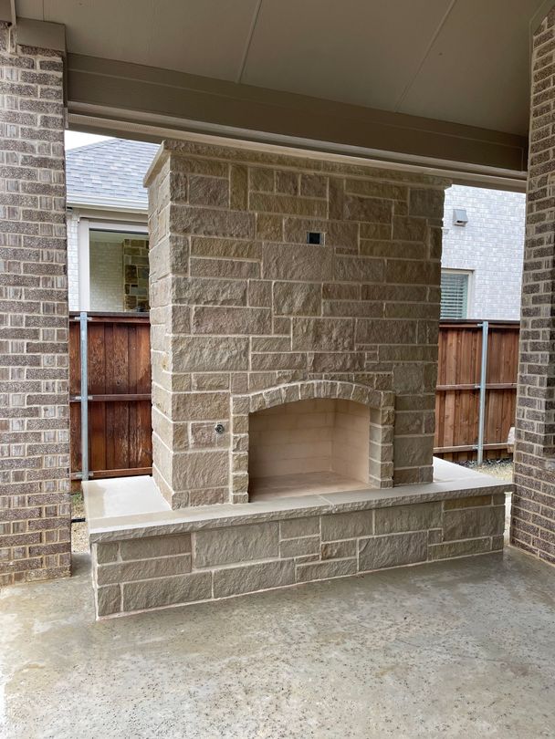 A covered outdoor patio featuring a stone fireplace, wicker furniture, and white curtains on a backyard lawn.
