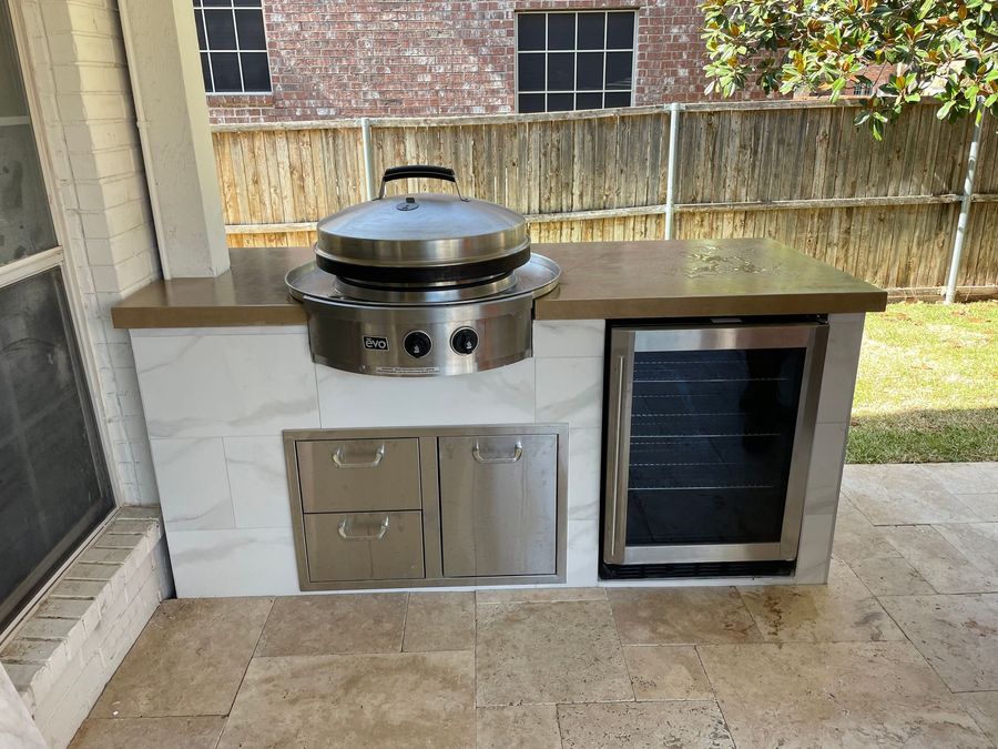 A patio with a stone outdoor kitchen, dining table with chairs, and pavers against a house with a tiled roof.