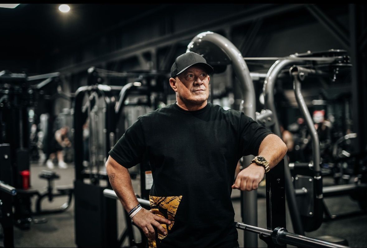 Man in black shirt and cap poses in gym, leaning on equipment, looking away.