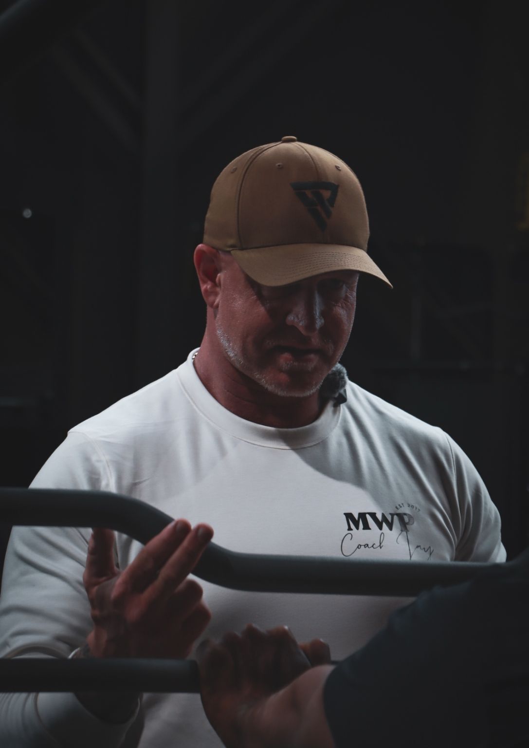 Man in cap and white shirt coaching with equipment, holding a bar. Dark setting.