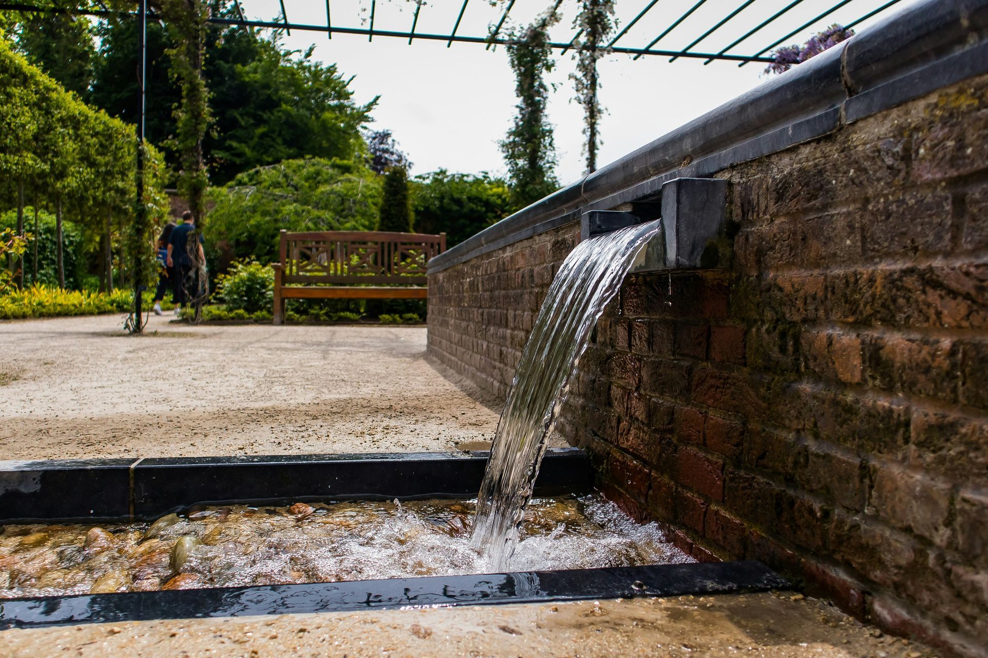 Water flowing from a brick wall into a shallow trough in a garden setting.