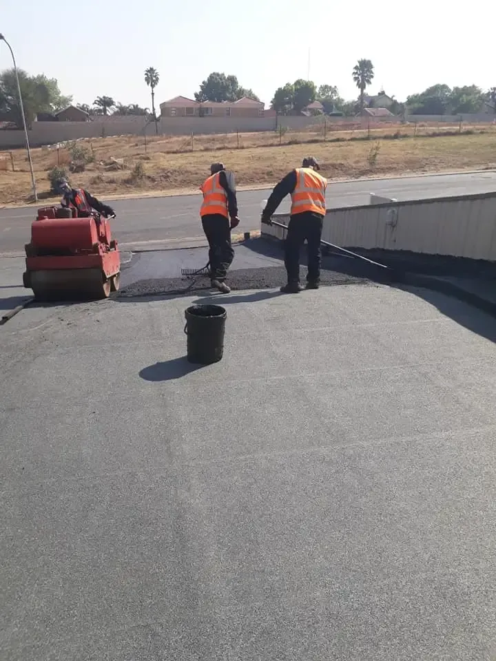 Asphalt paving: Two workers in orange vests lay asphalt, with a roller and a bucket. Sunny, outdoor setting.