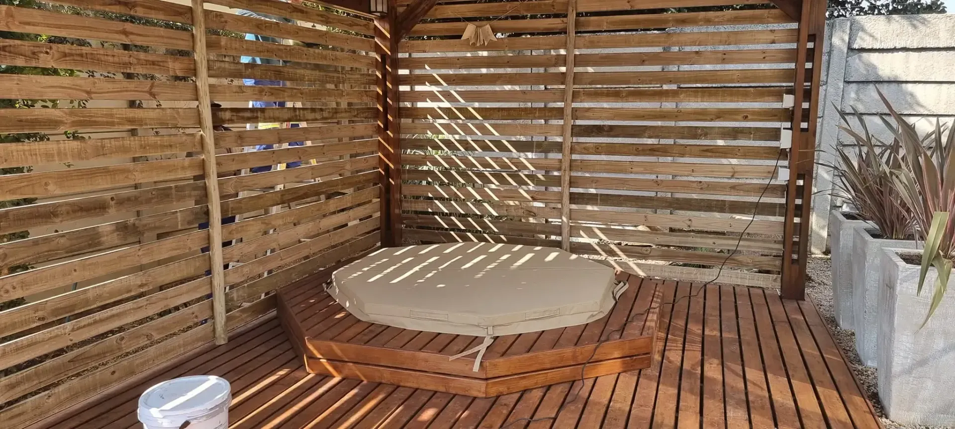 Wooden outdoor seating area with a cushioned round seat and slatted walls.