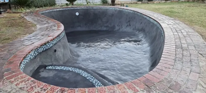 Empty, kidney-shaped swimming pool with brick border and gravel accents; outdoor setting.