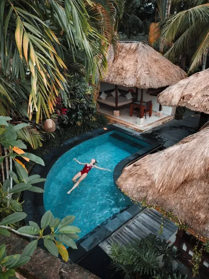 Woman floats in a blue pool surrounded by lush greenery, next to a thatched-roof gazebo.