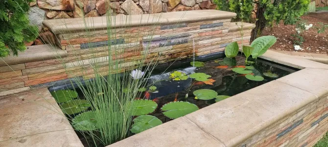 A rectangular stone pond with water lilies, fish, and plants, surrounded by a stone wall and a tiered stone backdrop.