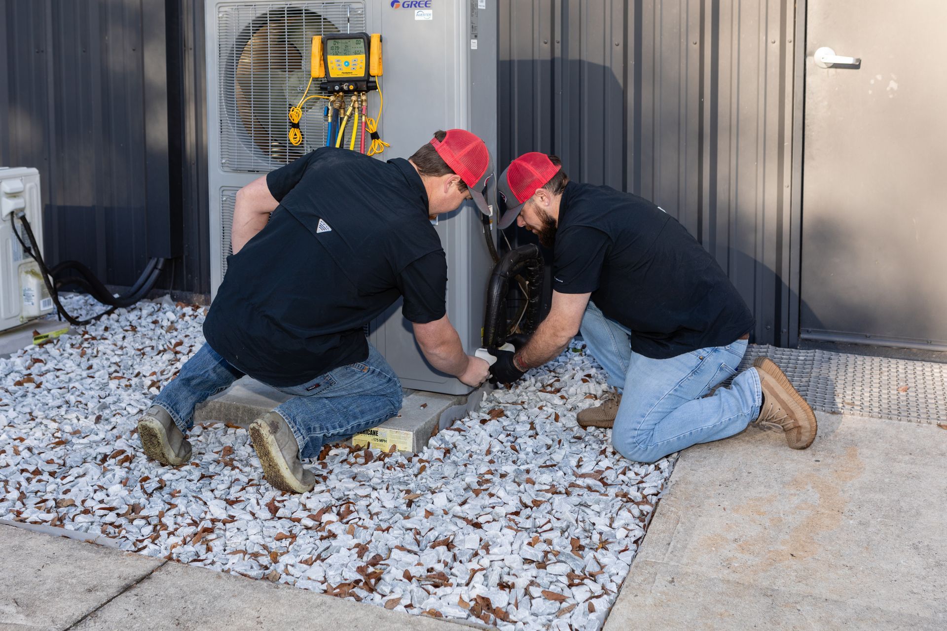 A technician in blue overalls tests an outdoor air conditioning unit with a multimeter.