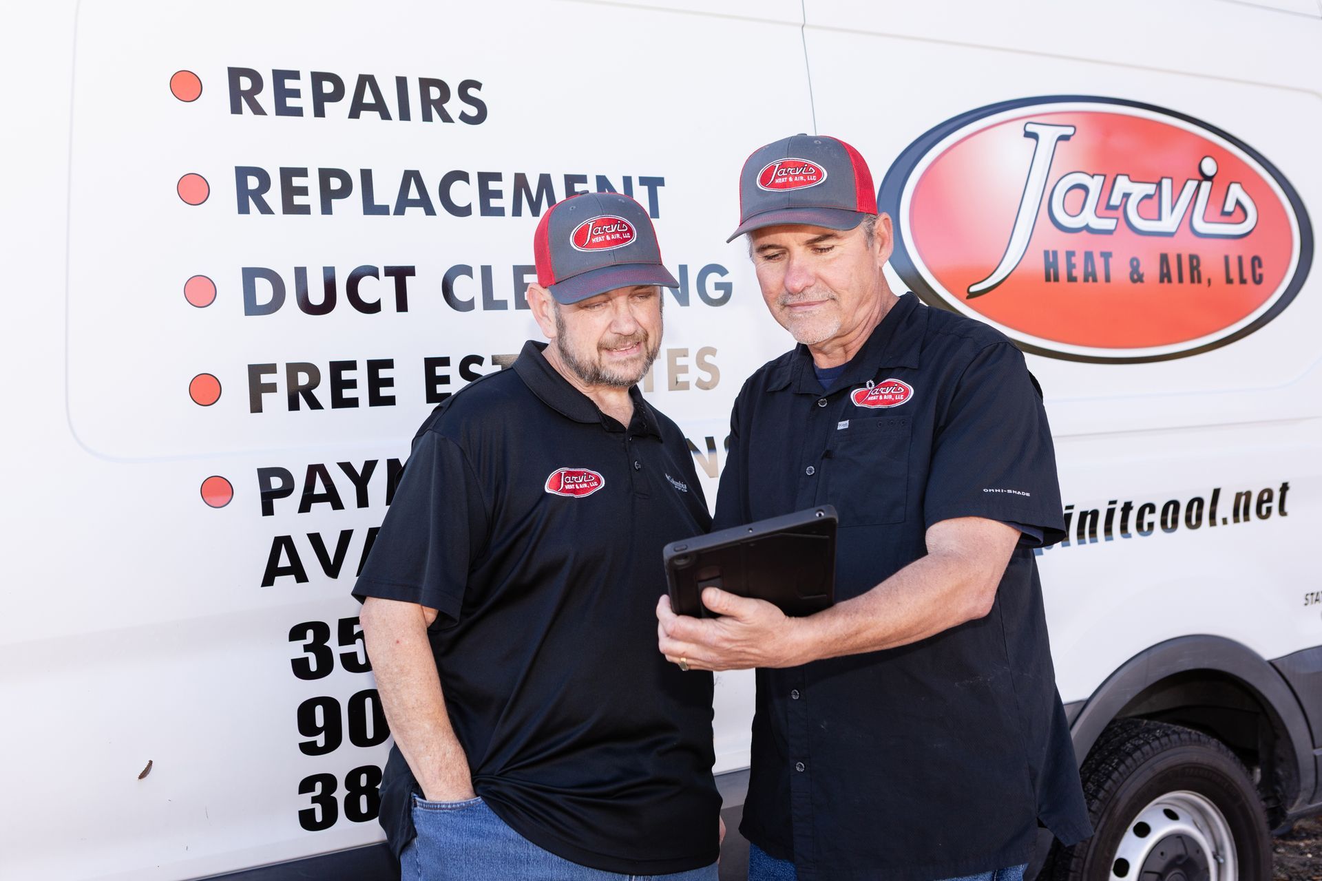 Two air conditioning units in a yard, near greenery and a brick wall.