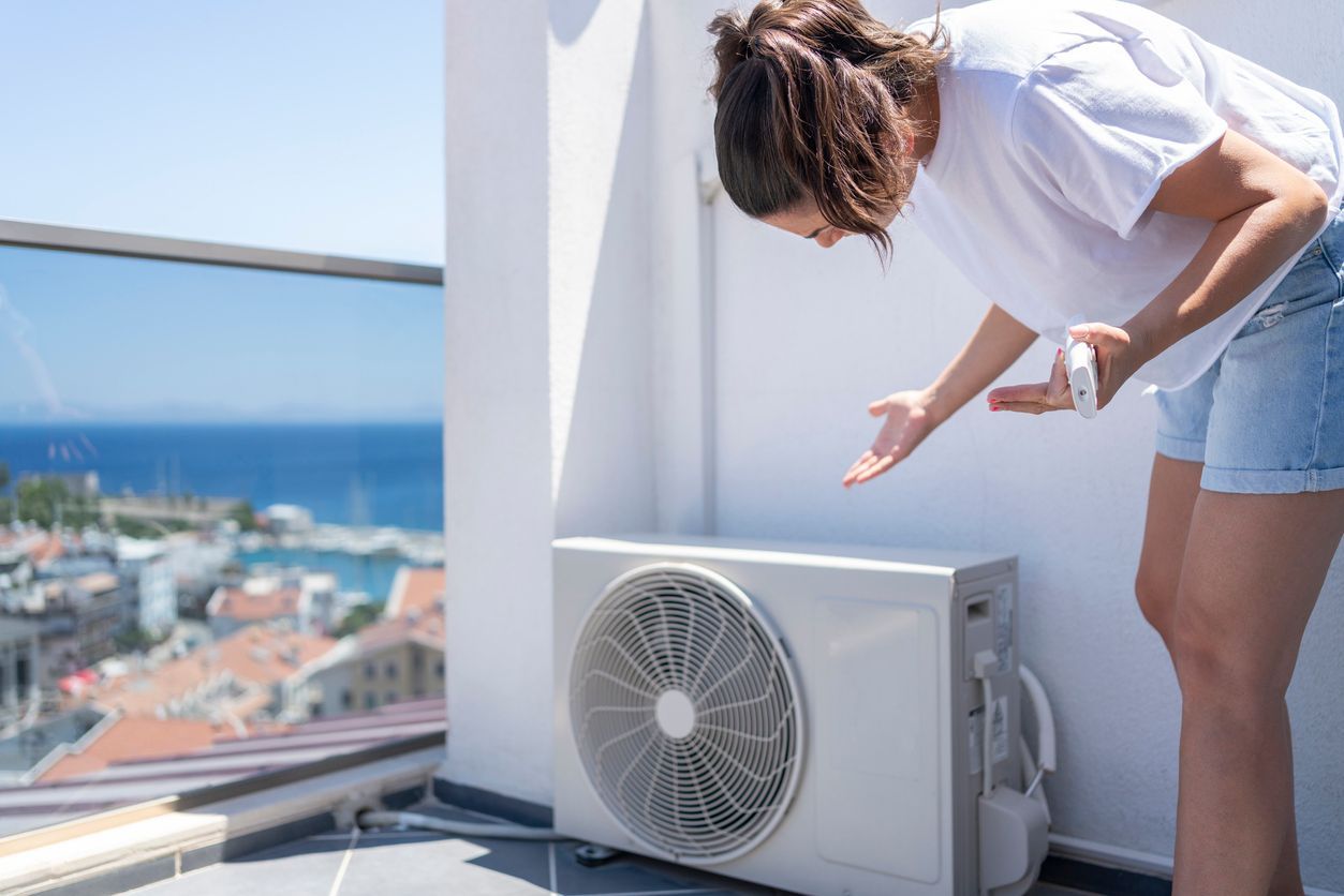 Woman examining an outdoor air conditioning unit on a balcony overlooking a coastal town.