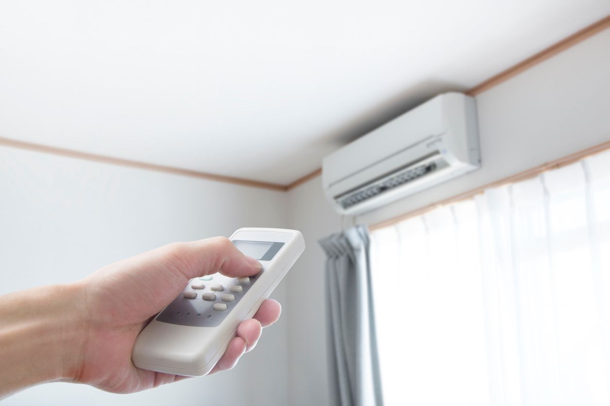 Hand holding a remote control pointing towards a mounted white air conditioner unit. Room interior.