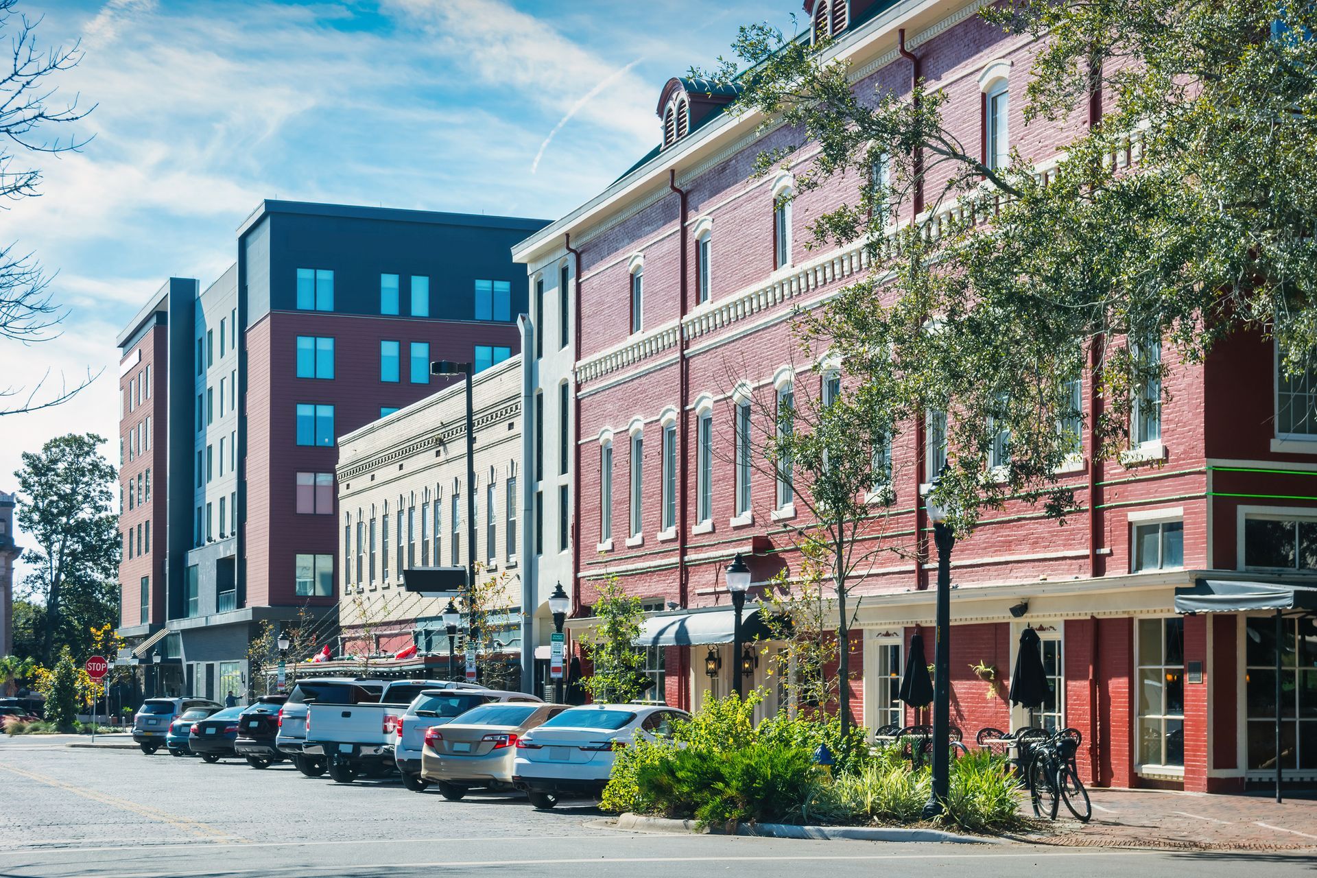 Street with parked cars, multi-story buildings of brick, white, and modern glass. Trees and blue sky.