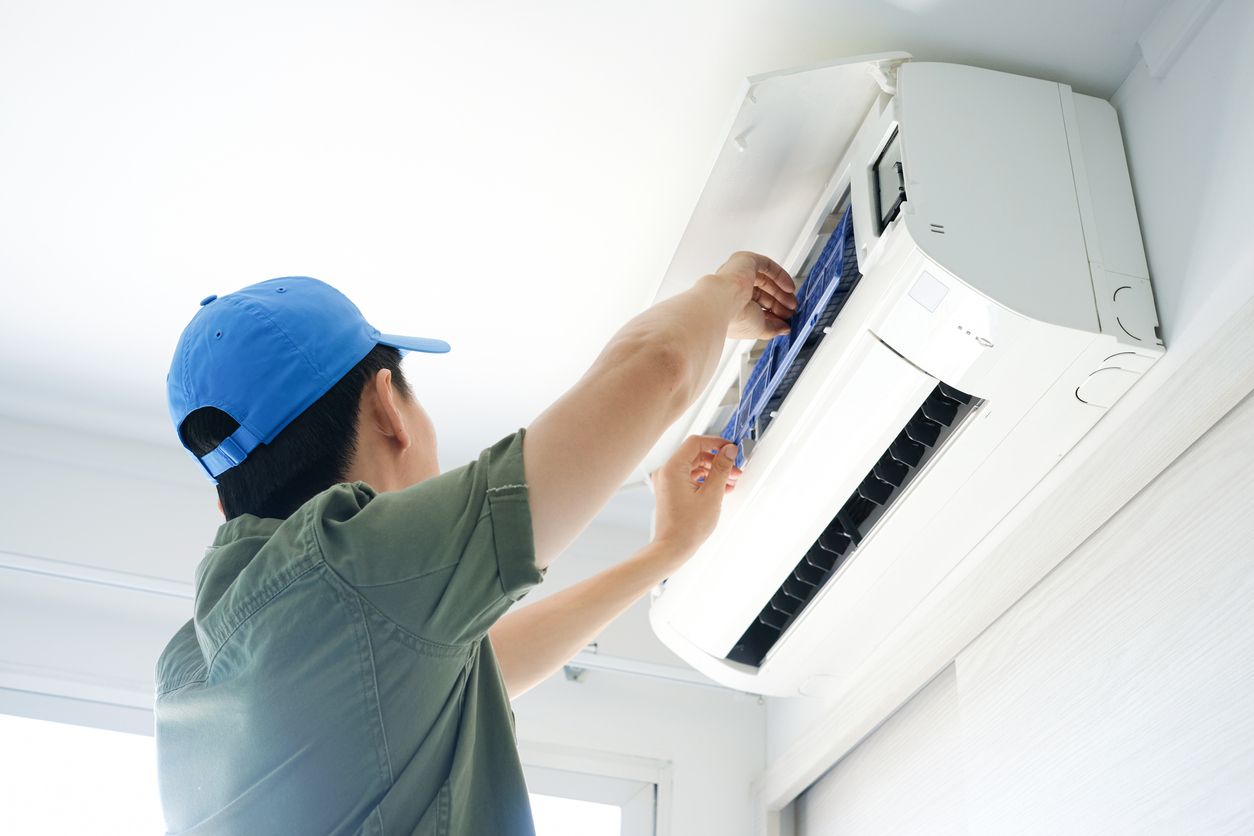 Person in a blue cap cleaning an air conditioner filter mounted on a white wall.